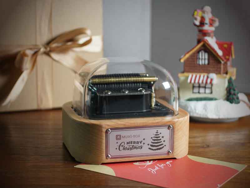 The Muro Box-N20 Standard programmable music box, fitted with a special Christmas-themed engraved nameplate. The scene is styled with holiday props: a Santa figurine climbing a small house and gift boxes tied with gold ribbons.