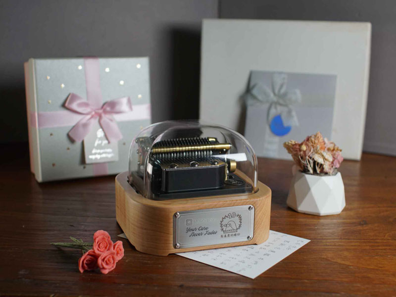 Muro Box-N20智慧音樂盒，客製化雷雕『Your Care Never Fades』與『有溫度的緣份』字樣以及綠繡眼圖案的金屬銘版，旁有小盆栽與木雕鳥，後方伴隨粉色絲帶禮物盒與乾燥花