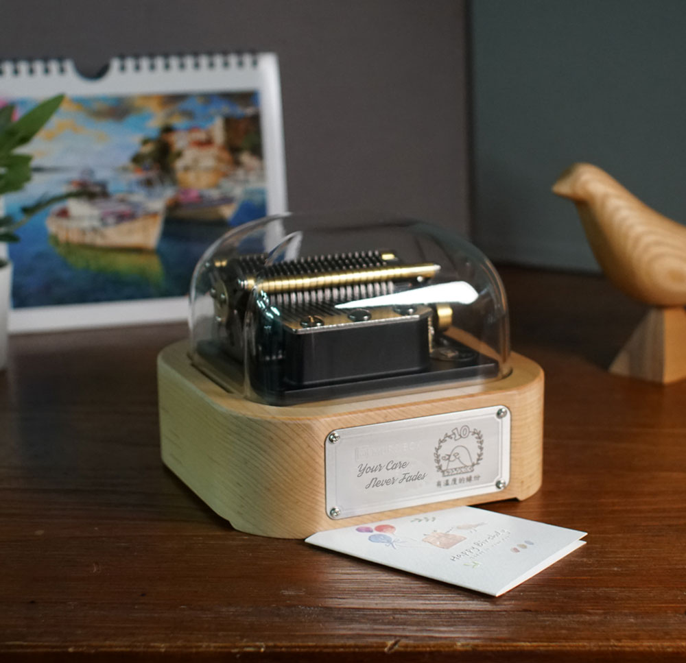 Muro Box-N20智慧音樂盒，客製化雷雕『Your Care Never Fades』與『有溫度的緣份』字樣以及綠繡眼圖案的金屬銘版，後方搭配小盆栽、風景月曆與木雕鳥裝飾