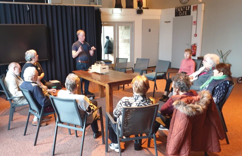 In the lounge of the nursing home, the elderly residents are seated in a semi-circle. A man (Joeri) is standing at the large table, attentively demonstrating and explaining the Muro Box-N20 smart music box placed on it.