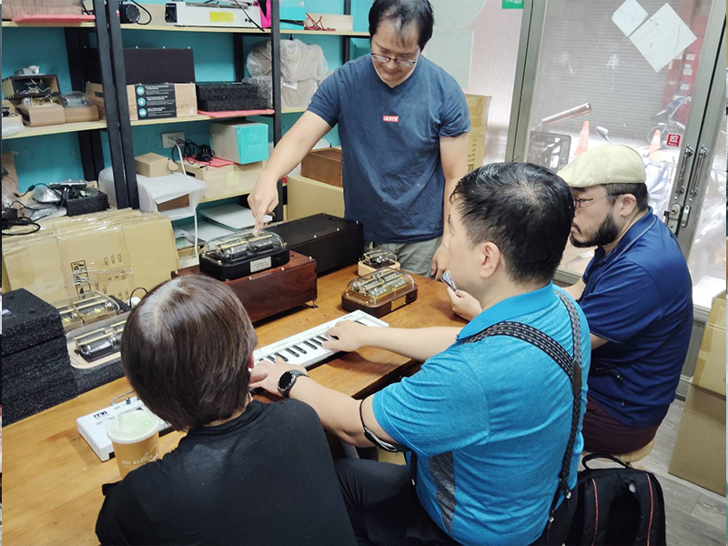 作曲家兼指揮家張俊彥與「Mysterium Musicum 音樂密契人聲室內樂集」的團員圍繞在桌前，共同專注研究智慧音樂盒 Muro Box-N40 穆風版。畫面上張老師正親自彈奏MIDI鍵盤來控制智慧音樂盒的同步演奏並仔細聆聽聲音與反應時間是否夠及時，團員們也一同觀賞 N40音樂盒雙機種的物理聲響與比對不同機種的音色