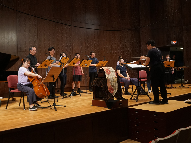 演出前的國家音樂廳舞台彩排現場。畫面中，「Mysterium Musicum 音樂密契人聲室內樂集」與樂手們正在台上準備，而舞台前方放置著一台智慧音樂盒 Muro Box-N40 穆風版，表面正蓋著一塊張俊彥老師特別挑選的精美花紋布料。此畫面記錄下作曲家兼指揮家張俊彥為了避免攝影機與聚光燈太早拍到音樂盒、提早破壞驚喜，特意將壓軸登場的 N40 遮蓋起來的貼心準備過程。