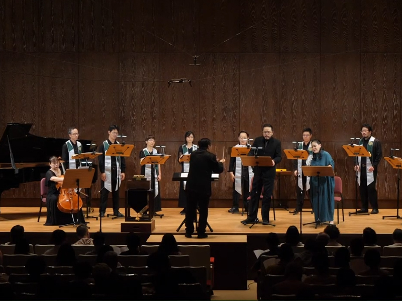 國家音樂廳的演奏會舞台中央，「Mysterium Musicum 音樂密契人聲室內樂集」與樂手們已經更換成正式登台服裝並各就各位，作曲家兼指揮家張俊彥站在指揮台中央，與身旁專屬舞台上放置的智慧音樂盒 Muro Box-N40 穆風版僅有一步之遙，便於張老師在需要時能輕鬆伸手觸碰正面旋鈕、啟動樂章尾聲的指定旋律。畫面同時記錄了懸掛在指揮家頭頂上方的黑色收音麥克風，展現現場針對歌唱者音量與各種樂器聲音大小所做的專業收音配置。